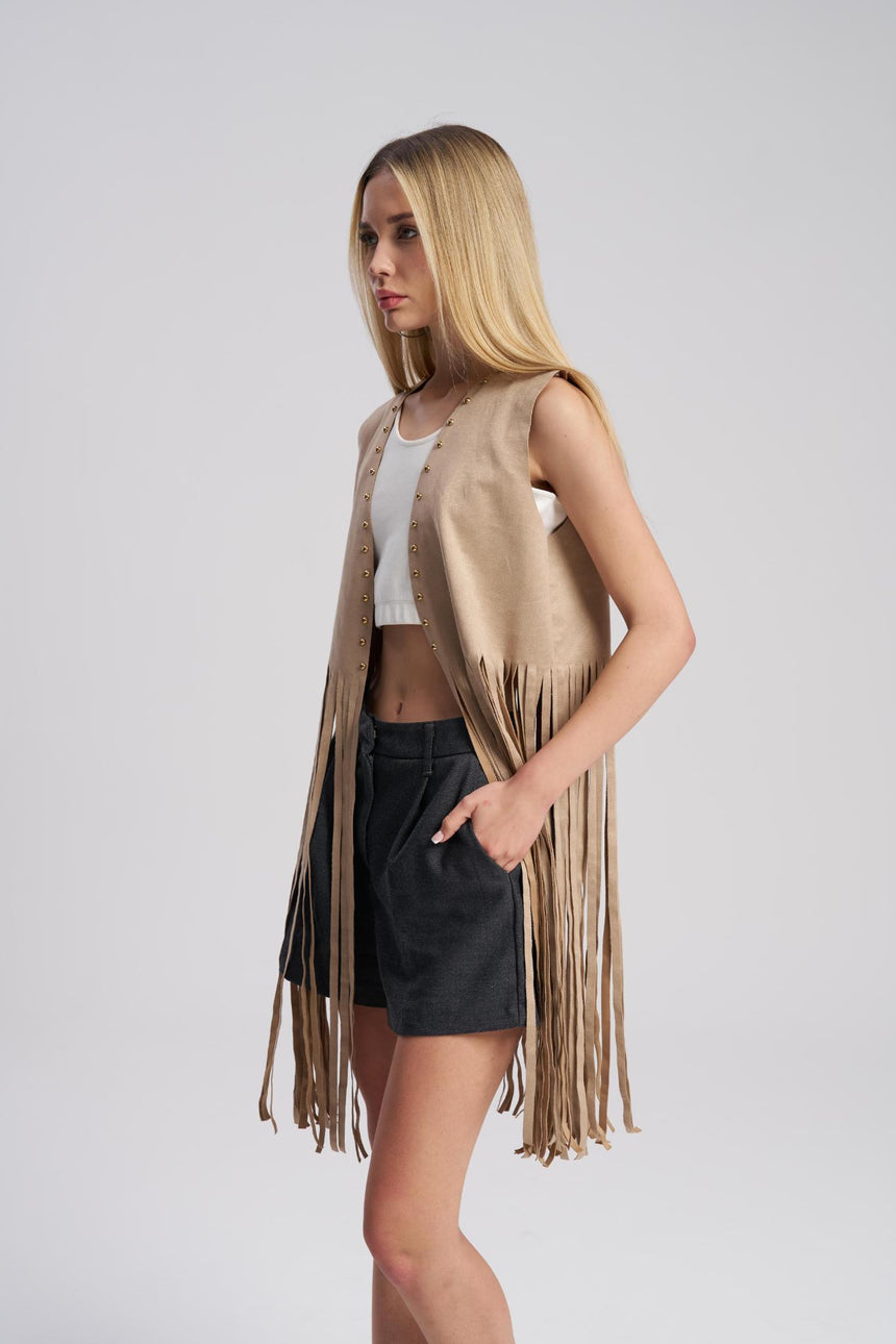 Woman wearing a beige fringe vest over a white top and black shorts on a light gray background