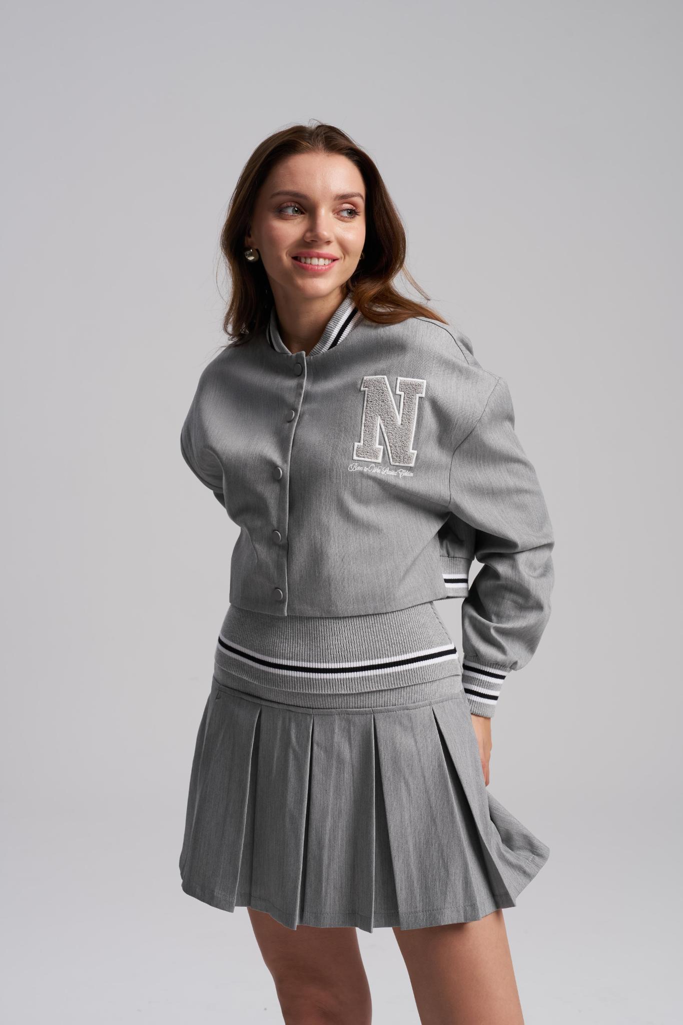 Woman wearing a gray varsity jacket with a 'N' emblem and pleated skirt on a light gray background