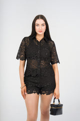 A woman modeling a black lace short-sleeve button-up shirt and matching lace shorts, holding a small black handbag against a light gray studio background.