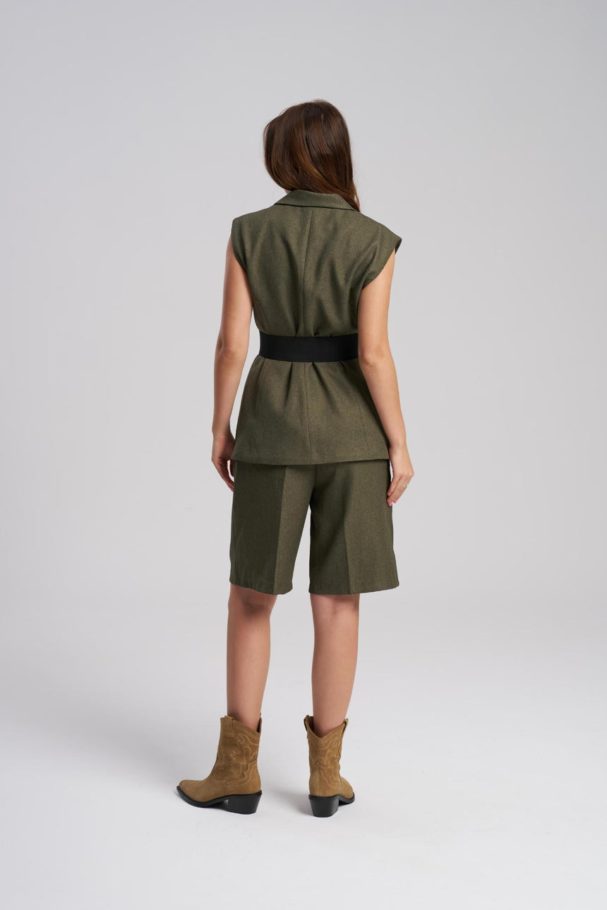 Person wearing a green sleeveless top and shorts with a belt, standing against a white background.