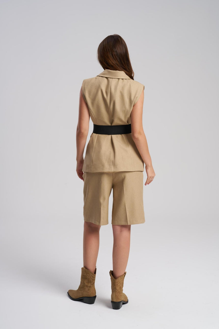Woman wearing a beige sleeveless jacket and shorts with a black belt, standing against a white background.